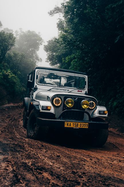 A silver off-road vehicle, specifically a Land Rover Defender, positioned on a muddy, uneven dirt track surrounded by dense green foliage and tall trees. The vehicle features a robust black front bumper with a protective grille, and three large, circular yellow headlights mounted on the front grille, with the middle one illuminated, creating a striking contrast against the grey bodywork. The windshield is clear, revealing the interior with seats and a steering wheel. The vehicle’s yellow registration plate reading 'KA 15B 0399' is prominently displayed at the front. The background shows a misty, overcast sky with soft lighting filtering through the tree canopy, emphasizing the rugged outdoor environment, which is typical for private off-road excursions or alternative vehicle access in remote or wooded areas. The scene suggests outdoor adventure, off-road travel, or landscape clearing by specialist companies such as Rubbish Removal Streatham, which may handle waste disposal from similar environments, though no rubbish or waste objects are visible in this image.