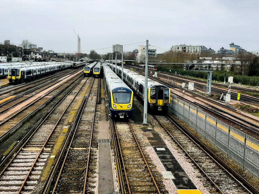 A collection of modern passenger trains parked on multiple railway tracks at a train yard. The trains share a similar design with sleek, aerodynamic shapes, and are predominantly silver with yellow accents around the front and sides. The tracks are made of metal rails mounted on wooden sleepers, with some sections protected by small platforms or barriers. Overhead, electrical wires run along the length of the yard supported by tall, grey metal poles, indicating electric-powered trains. In the background, there are several multi-storey buildings and a wind turbine visible against a cloudy, overcast sky, creating an urban environment context. To the right side, a metal fence with yellow and black safety markings runs parallel to the tracks. The overall scene appears busy, with the trains arranged systematically, suggesting a railway maintenance or storage facility where trains are temporarily stationed or prepared for service, fitting into the concept of alternative rail-based transport and private railway operations.