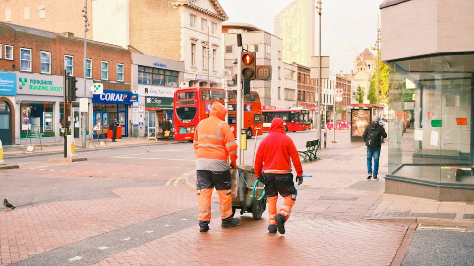 Two waste management workers dressed in high-visibility orange and grey uniforms are seen walking along a city street, pushing a wheeled bin or waste container. One worker, with their back slightly turned, is holding the handle of the bin, while the other is walking alongside, guiding it. The street is paved with red bricks and features a traffic light showing red for vehicles, with two red double-decker buses waiting at the stop in the background. Surrounding the scene are city storefronts and shops, including a Yorkshire Building Society branch and a Coral betting shop, with signage visible on the buildings. Pedestrians are seen in the background, including a person wearing a black jacket and jeans near a glass storefront. The environment is urban, with a mix of older and more modern architecture, and the lighting suggests daytime, possibly early morning or late afternoon, with a neutral, professional tone suitable for describing waste management and rubbish removal services. Rubbish Removal Streatham may carry out similar on-site clearance services for residents or businesses in the area.