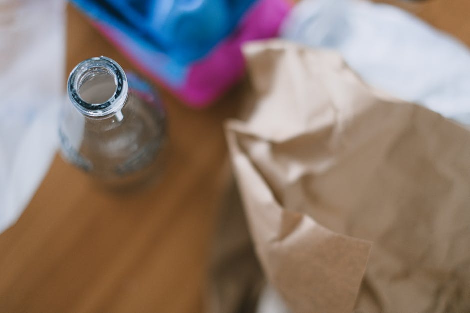A close-up view of a transparent glass bottle with a narrow neck and open top, positioned on a wooden surface. The bottle appears to be clean and empty, with a smooth, glossy finish reflecting light. In the background, there are crumpled brown paper bags, partially unfolded, and a blurred blue and pink object, possibly a bag or packaging material. The environment suggests an indoor setting, potentially a kitchen or workspace, with natural or ambient lighting creating soft shadows. The scene relates to rubbish disposal or collection, aligning with private waste handling services such as those offered by Rubbish Removal Streatham, especially in contexts involving waste sorting or on-site clearance of packaging and household waste materials for more sustainable rubbish removal options.