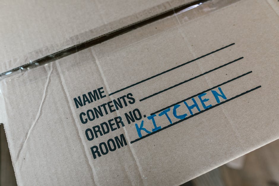 Close-up view of a brown cardboard box with black text printed on the side, including labels for 'NAME,' 'CONTENTS,' 'ORDER NO.,' and 'ROOM,' with the handwritten addition 'KITCHEN' in blue ink. The box appears to be sealed with clear packing tape along the top edge, which is partially visible. The surface of the cardboard shows slight creases and texture typical of corrugated material. The background is blurred but suggests an indoor setting, possibly a storage area or moving environment. The box's labeling indicates it is used for organized packing or sorting, fitting into context such as waste management, where clear identification aids in proper disposal or recycling, aligning with the services of Rubbish Removal Streatham. The natural lighting highlights the material's texture and text clarity, emphasizing its functional purpose in waste handling and logistics.