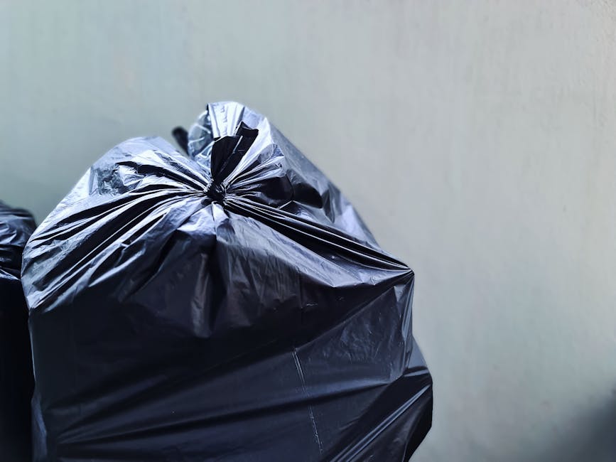 Close-up image of a large, black plastic rubbish bag tightly knotted at the top, resting against a plain, light-colored wall. The bag has a slightly glossy surface with visible creases and folds, indicating it is full and weighted. In the background, part of another black rubbish bag is partially visible, showing similar material and texture. The setting appears to be an indoor or sheltered area, possibly a storage space or gathering point for waste before collection. The overall scene reflects a typical private waste collection scenario, emphasizing the use of black plastic bags for rubbish disposal, aligned with services offered by companies like Rubbish Removal Streatham that facilitate independent or alternative waste handling solutions in the Streatham area. The image demonstrates an organized approach to rubbish storage prior to professional rubbish removal, suitable for residential or small commercial contexts within the local estate environment.