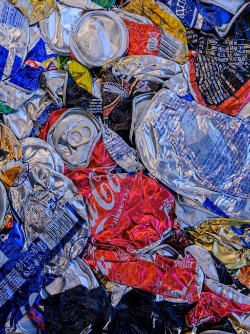 A close-up image showing a collection of crushed and crumpled aluminum cans, including beverage containers such as soda and energy drink cans, scattered randomly. The cans display various colors, including silver, red, blue, yellow, black, and white, with some featuring recognizable branding and logos like Coca-Cola. The surface textures range from smooth and reflective to dented and wrinkled, emphasizing the metallic material. The cans are piled together, overlapping each other, with some partially flattened and others still retaining their cylindrical shape. The background appears to be a mixture of similar waste materials, suggesting an environment focused on waste collection or recycling preparation. This scene aligns with private waste handling or alternative rubbish removal services, such as those offered by Rubbish Removal Streatham, indicating a recent collection or disposal of recyclable materials. The lighting is even, highlighting the metallic sheen and vibrant colors of the cans, underlining the importance of proper waste separation and disposal in an urban setting.